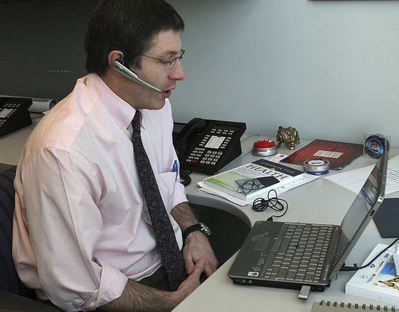 A man with his sleeves rolled up speaks into a headset while staring at a laptop.